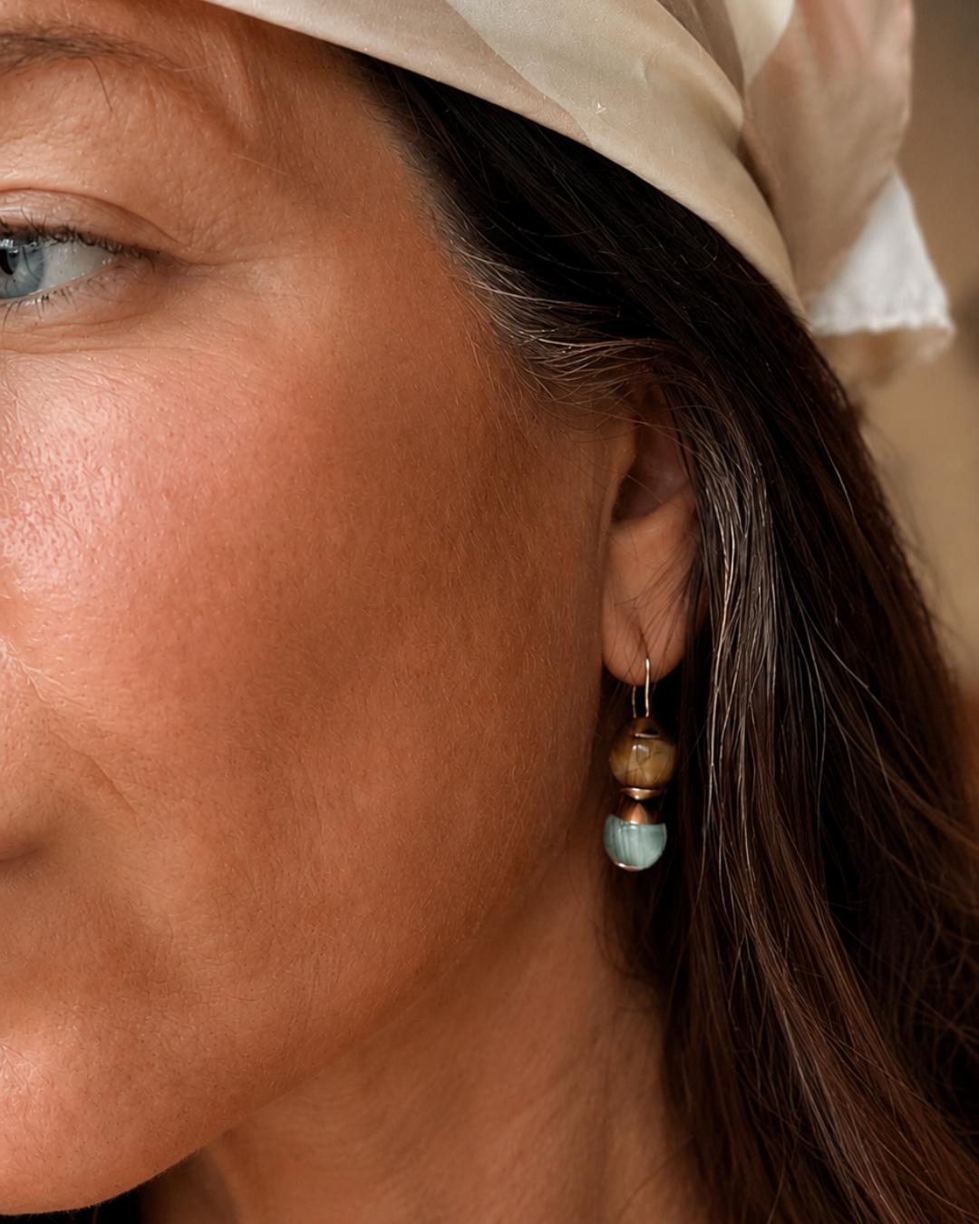 A close-up of a woman's ear wearing a delicate gold dangle earring with a brown wooden bead, a light blue stone bead, and a small turquoise bead.