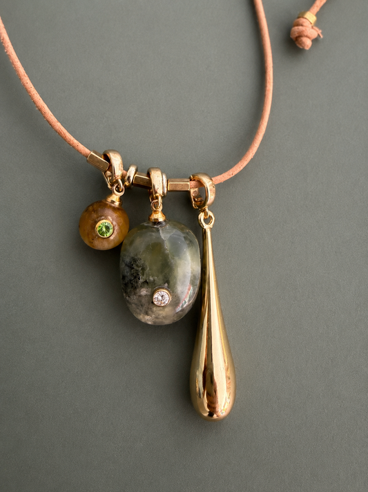 A gold-toned necklace with a peach cord, featuring a triple-hanging pendant design: a small brown bead with a green stone, a polished gray stone with a clear crystal inset, and a teardrop-shaped gold pendant, shown on a dark gray background.