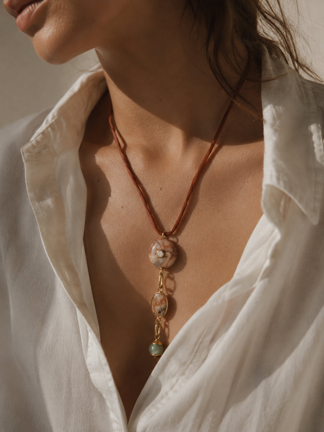 A close-up of a woman wearing a bohemian-style necklace with a brown cord and a pendant made of marbled stone and gold-toned metal, styled with an open white shirt.