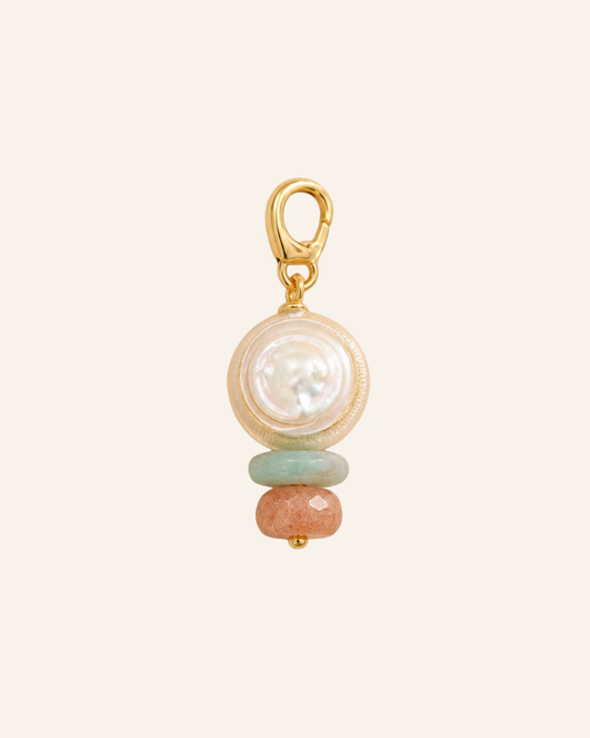 A gold charm featuring a round white pearl, a light green stone bead, and a peach-colored stone bead, set against a solid pale cream background.