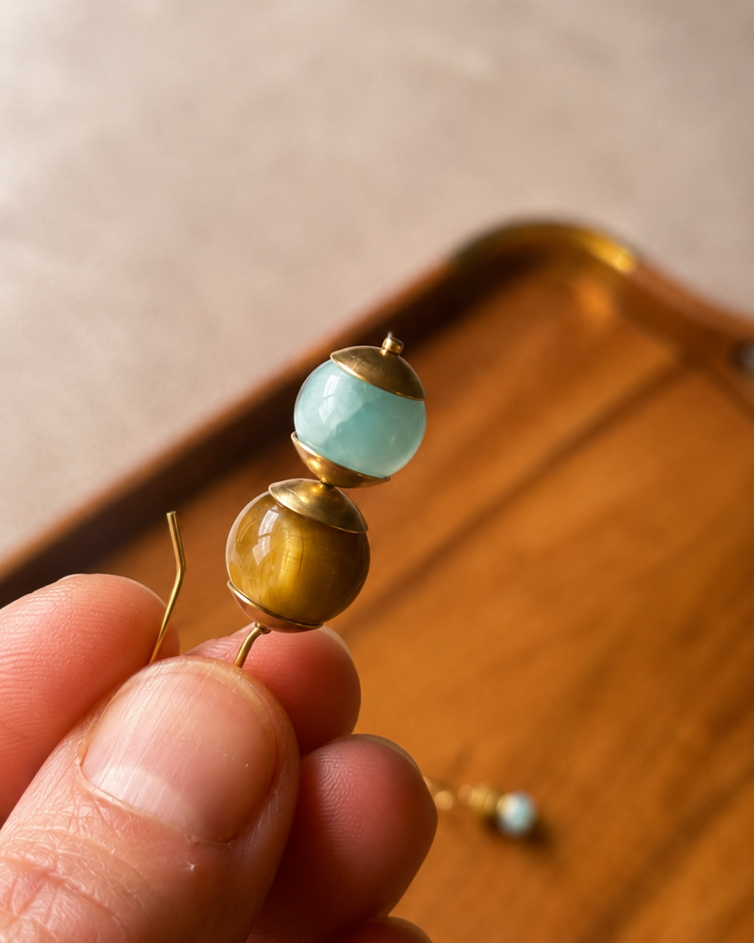 A close-up of a hand holding a gold-toned beaded earring with a light blue-green round bead and a smaller gold-toned bead, with a wooden tray blurred in the background.