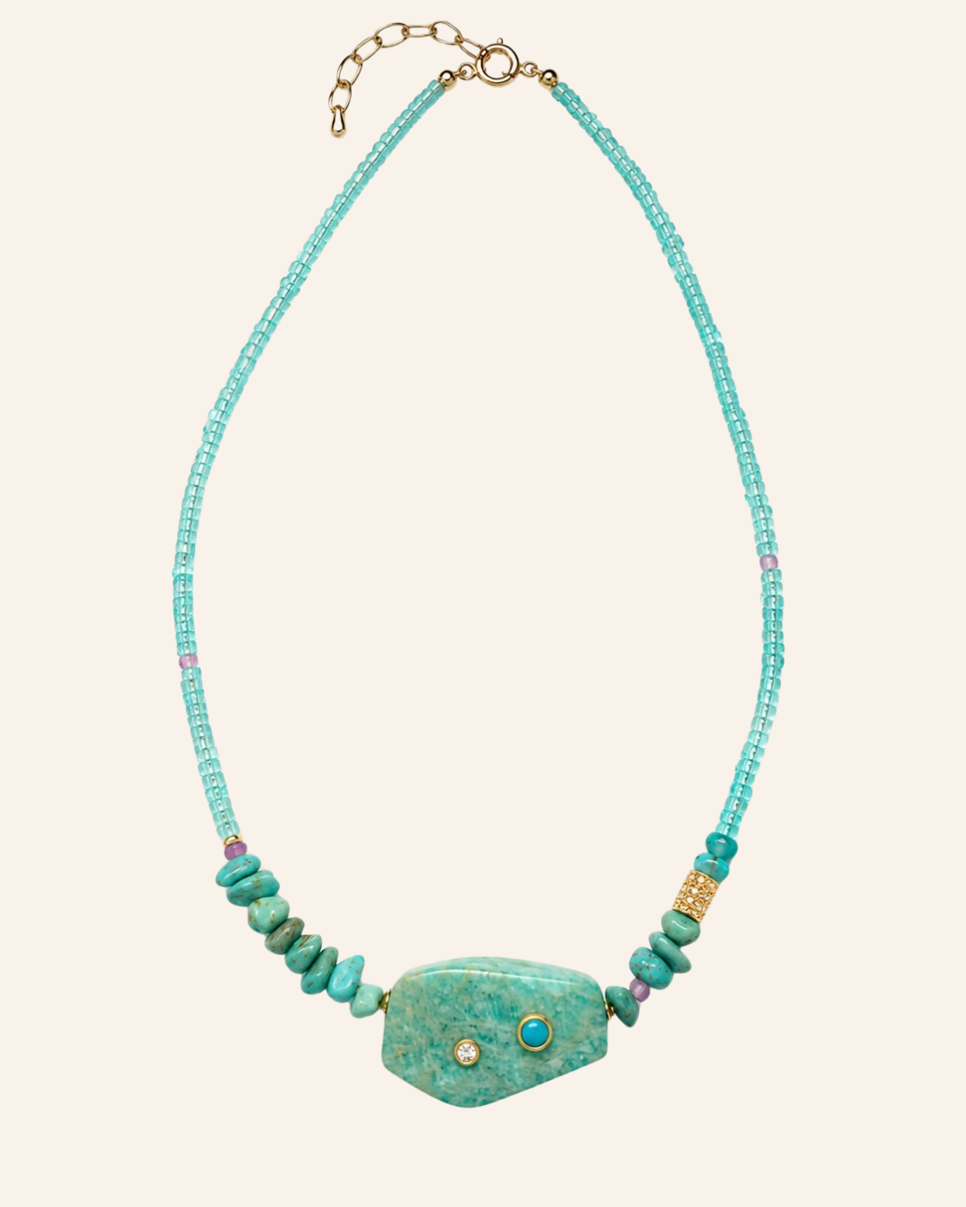 A turquoise and green stone beaded necklace with a large central turquoise stone pendant, gold chain extender, and small pink accent beads, shown on a light beige background.