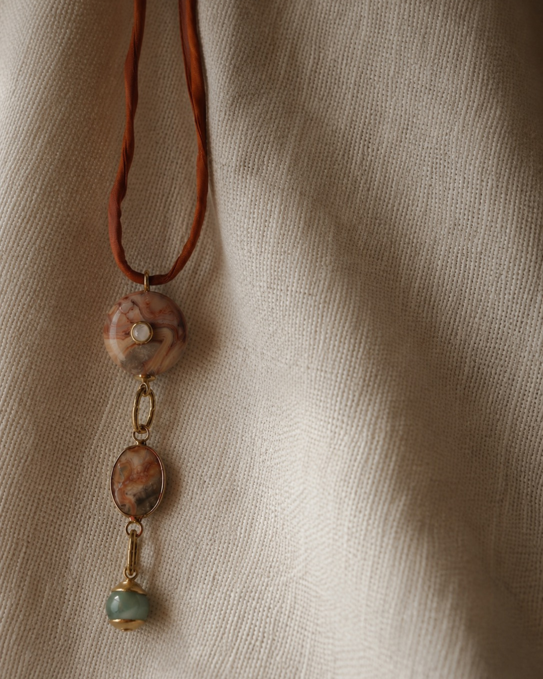 A bohemian-style pendant necklace featuring a brown cord, a round stone bead with a central pearl, an oval stone bead, and a dangling light blue stone charm with gold-toned metal accents, displayed on a textured beige linen fabric.