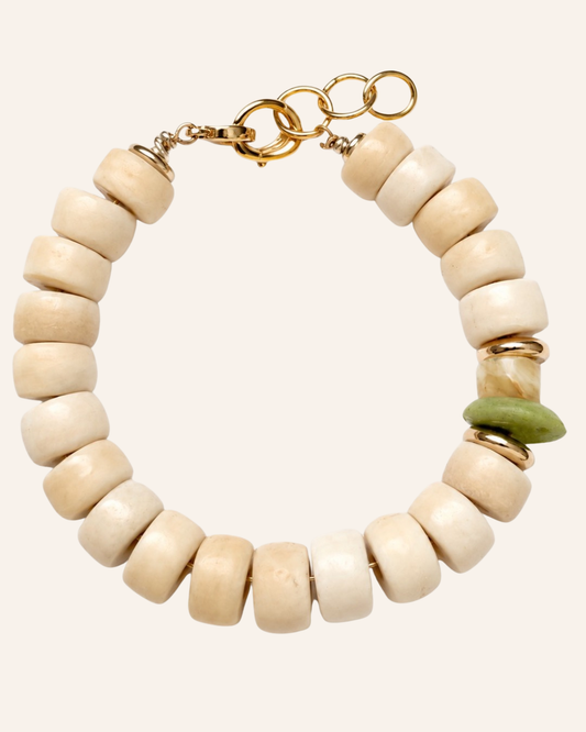 A beaded bracelet featuring matte cream-colored disc beads, gold-toned metal rings and clasps, and a single green stone accent bead, shown on a solid pale beige background.