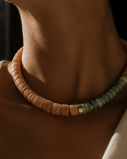 A close-up of a person's neck and collarbone wearing a chunky beaded necklace. The necklace features light tan cylindrical beads on one side and light green oval beads on the other, separated by small gold spacer beads.