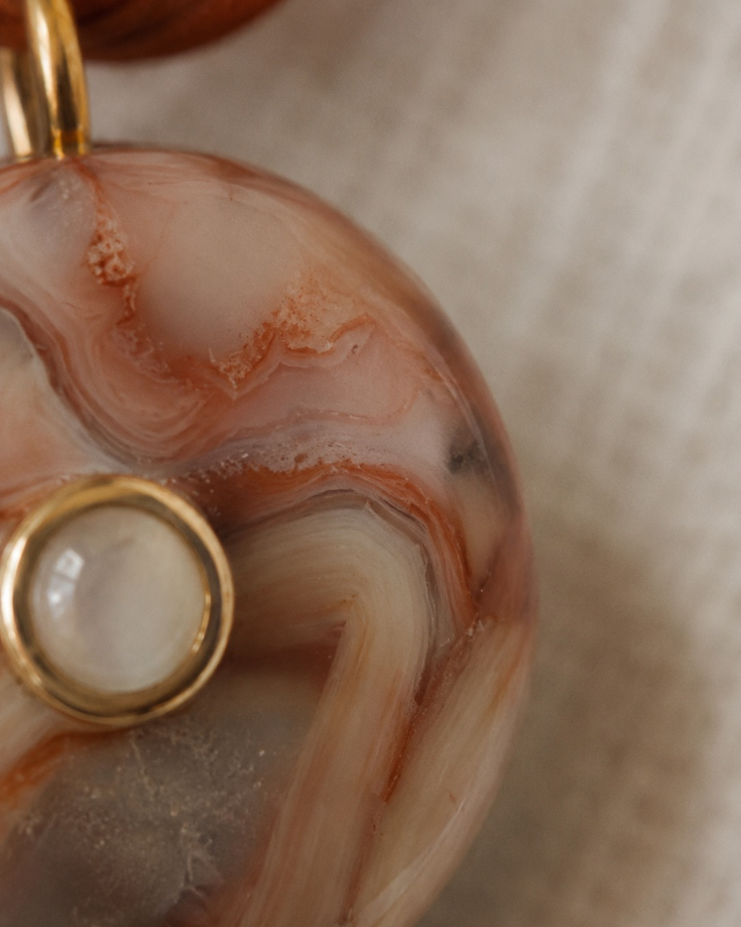 Close-up of a round agate pendant with swirling peach, cream, and brown natural stone patterns, featuring a gold bail and a gold bezel-set moonstone accent.