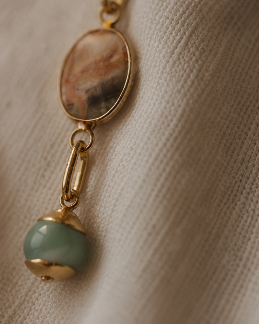 A close-up of a gold-toned pendant necklace featuring an oval marbled stone and a dangling round light green bead charm, resting on a textured beige fabric.