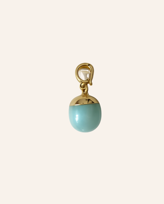 A small jewelry charm or pendant with a glossy light aqua oval drop and polished gold-tone metal top and loop, centered on a plain warm off-white background.
