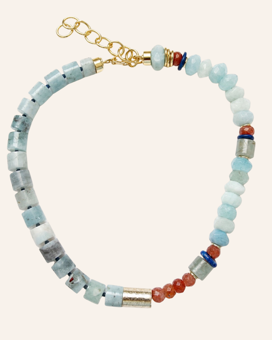 A beaded necklace featuring light blue and white stone beads, small red-orange beads, silver-toned cylindrical spacers, and a gold-toned chain clasp, displayed on a solid pale pink background.