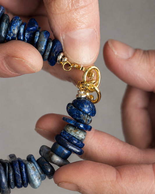 Close-up of a beaded necklace with dark blue lapis lazuli-style round beads and a gold clasp, held by two hands against a neutral gray background.