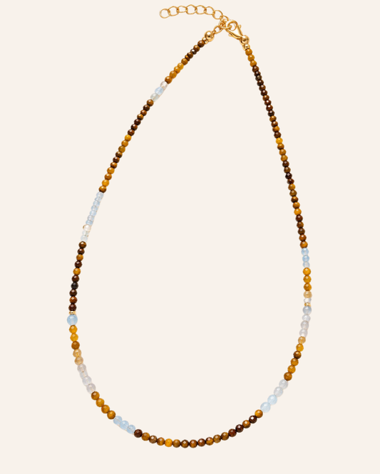 A beaded necklace with alternating strands of brown and amber-yellow beads, light blue beads, and small gold spacer beads, finished with a gold clasp on a light beige background.