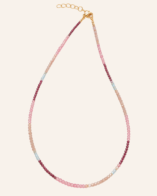 A delicate beaded necklace with a gold clasp and extender chain, featuring beads in pink, light blue, and dark red, displayed on a solid cream background.