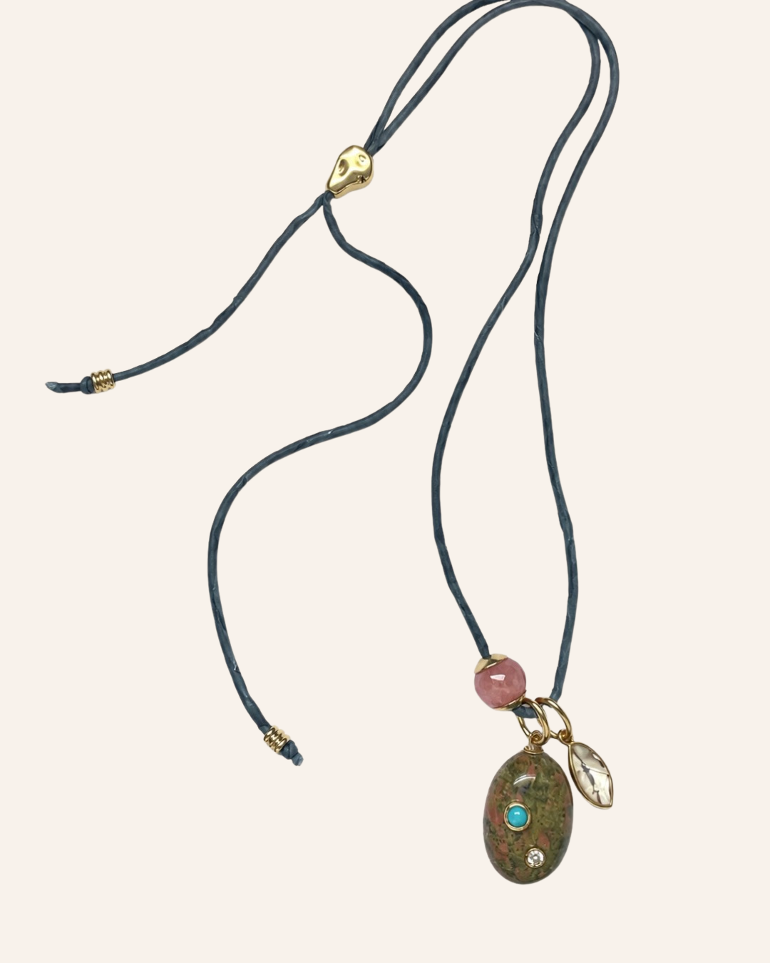 A bohemian-style necklace featuring a dark teal cord, a gold-toned adjustable slider, a pink bead, and a large oval stone pendant with a turquoise and clear crystal accent.