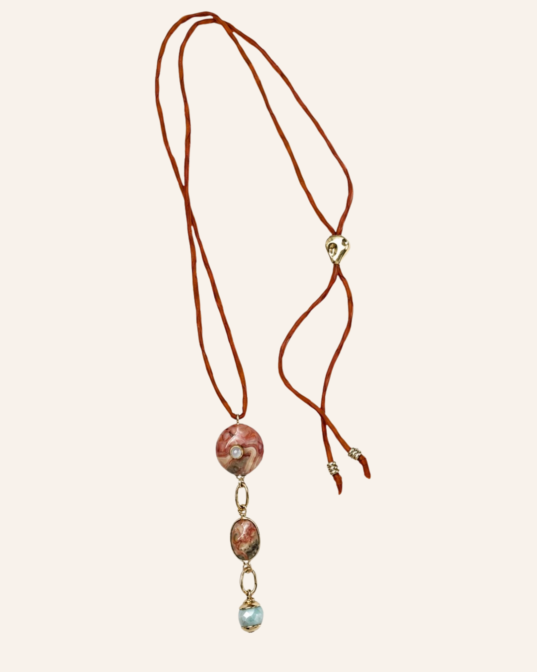 A bohemian-style necklace featuring a rust-brown cord, three marbled pink and brown beads, and a dangling light blue bead, shown against a solid pale pink background.