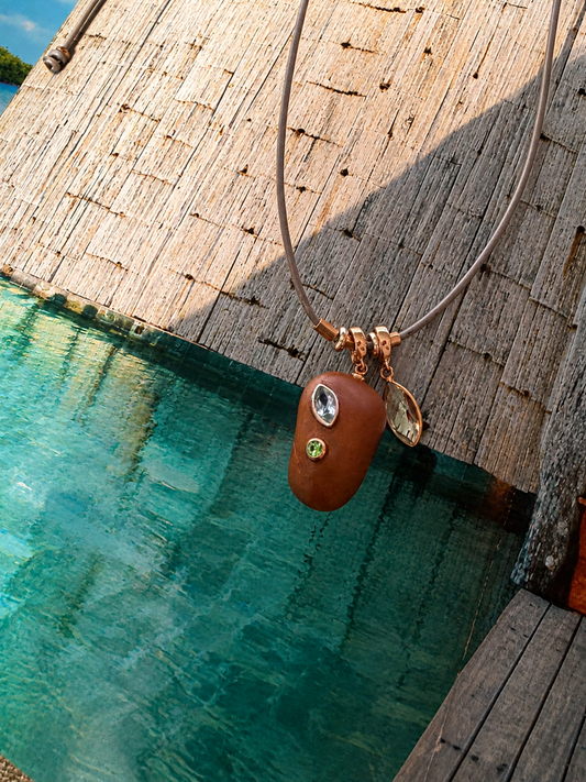 A brown leather necklace with a polished brown pendant featuring a clear oval stone and a small green stone, hanging over turquoise water next to a wooden dock.