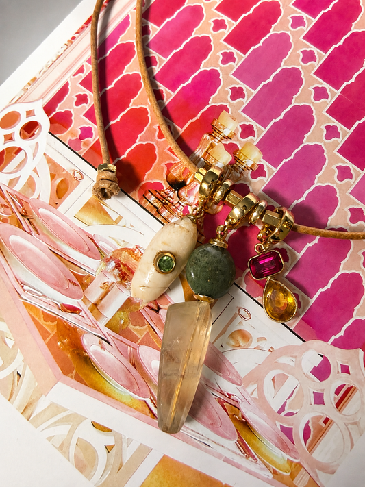 A bohemian-style pendant necklace with a brown cord, gold-toned beads, a cream-colored stone, a green stone, and a large clear crystal point, displayed on a pink patterned background.