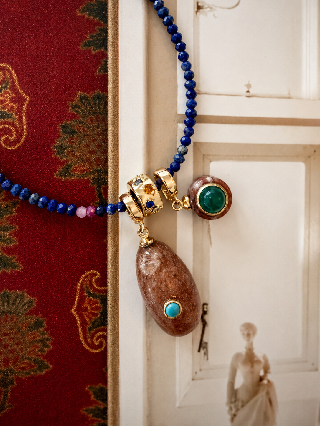 A close-up of a necklace draped over a white door, featuring deep blue beads, gold-toned hardware, a large brown stone pendant with turquoise and green stone accents, and a smaller round green stone charm.