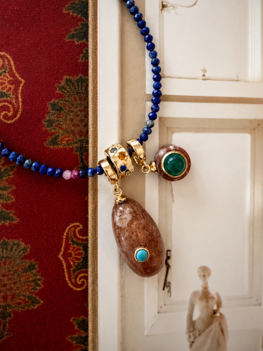 A close-up of a necklace draped over a white door, featuring deep blue beads, gold-toned hardware, a large brown stone pendant with turquoise and green stone accents, and a smaller round green stone charm.