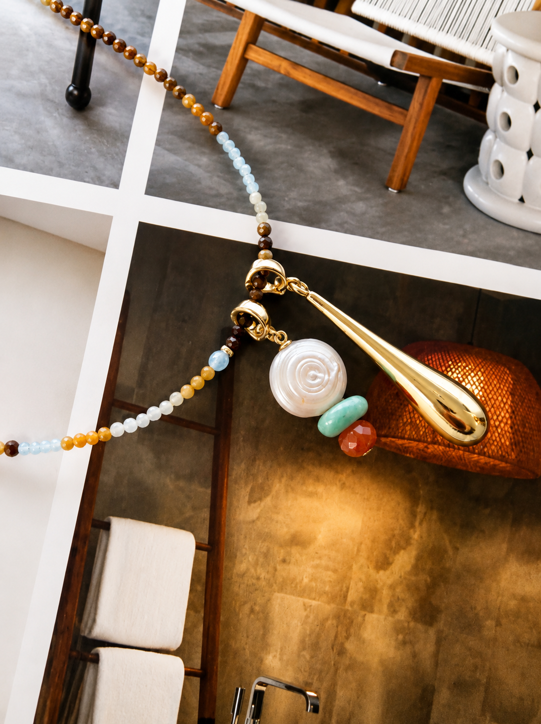 A bohemian-style beaded necklace with a large gold teardrop pendant, a white rose charm, and small turquoise and red beads, displayed in a modern bathroom setting.