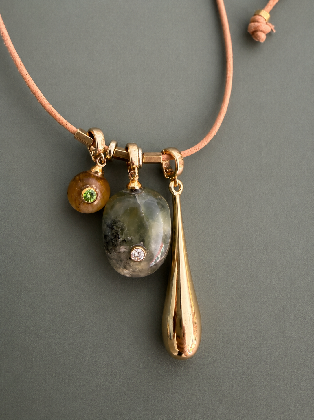 A gold-toned necklace with a peach cord, featuring a triple-hanging pendant design: a small brown bead with a green stone, a polished gray stone with a clear crystal inset, and a teardrop-shaped gold pendant, shown on a dark gray background.