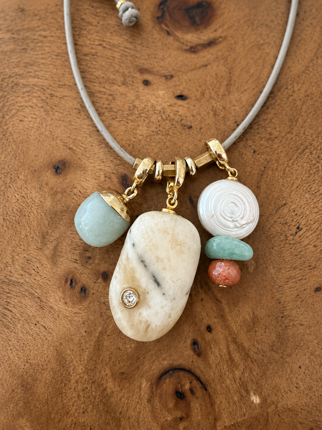 A beaded necklace with a gray cord and multiple stone charms, including a large cream-colored stone, a light blue stone, a white carved spiral bead, and a small coral bead, displayed on a wooden surface.
