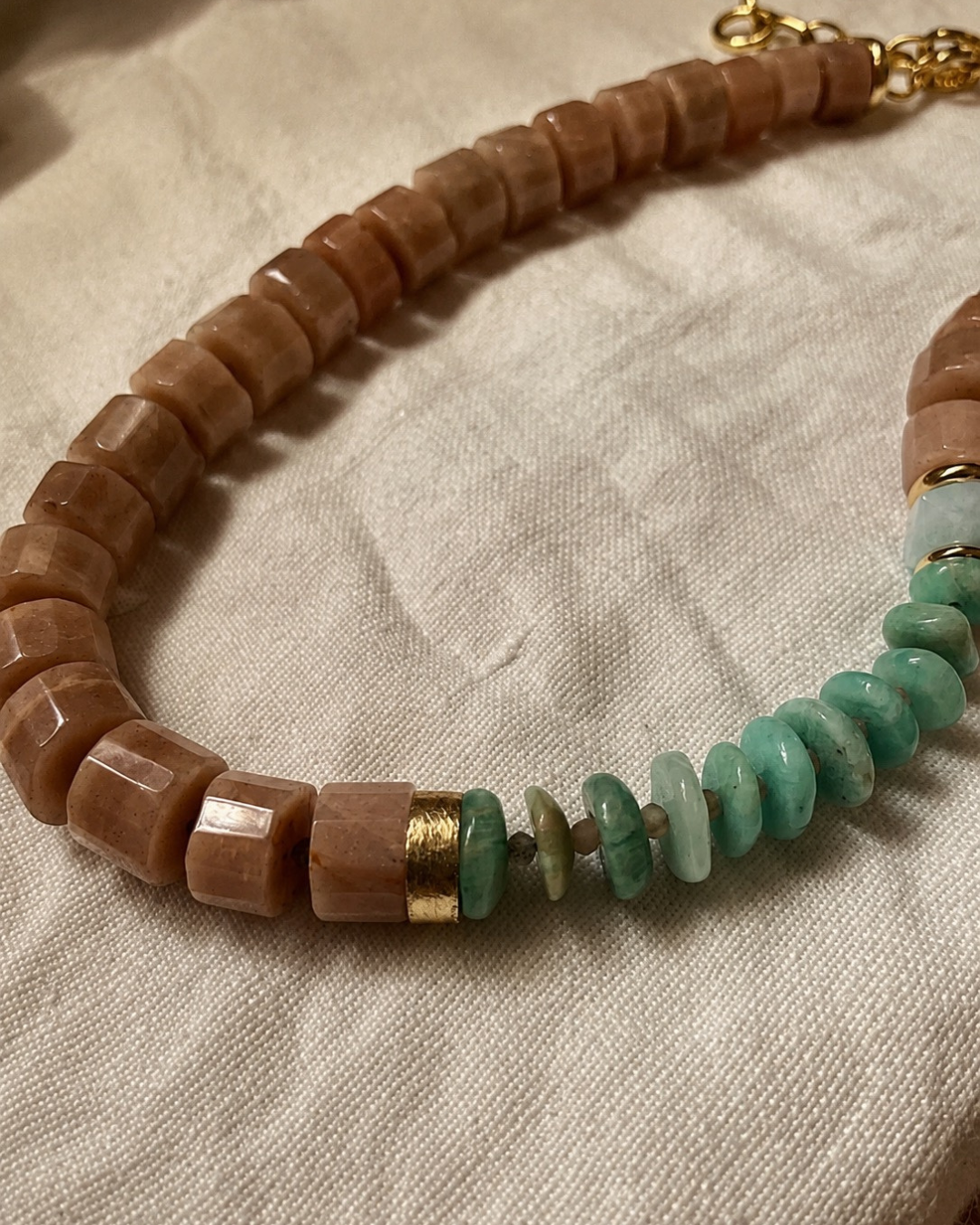 A beaded necklace made of faceted light brown stone beads, turquoise green stone disc beads, and small gold spacer rings, laid on a textured off-white fabric background.