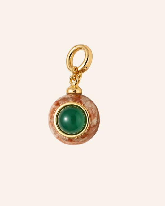 A round pendant charm featuring a polished green stone in the center, surrounded by a gold bezel and a textured brown and white marbled outer ring, with a gold hanging loop at the top, shown on a solid pale beige background.