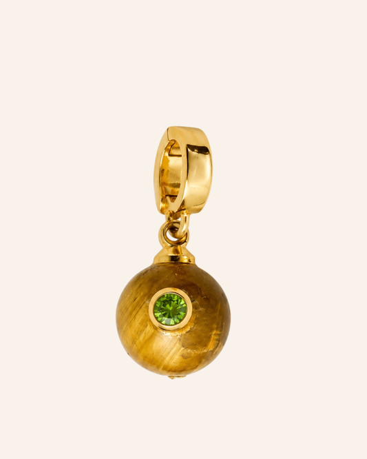 A gold huggie hoop earring with a dangling spherical charm featuring a green gemstone, shown on a solid pale cream background.
