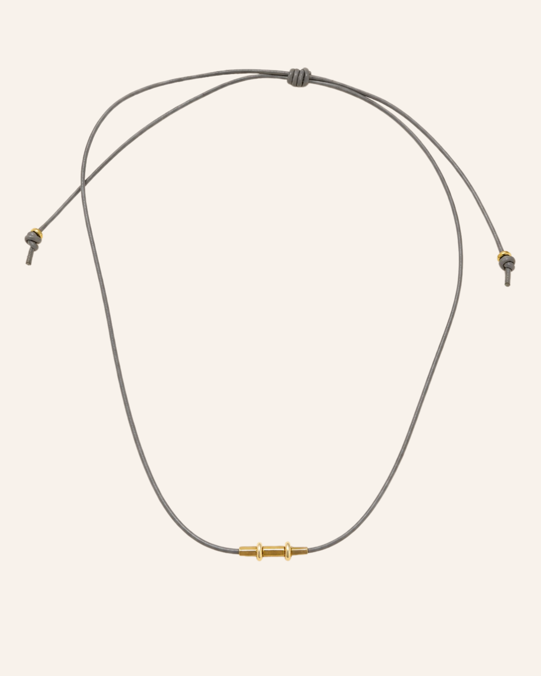 A minimalist adjustable cord necklace with a central gold-toned bead and gold-toned toggle details, displayed on a solid pale pink background.