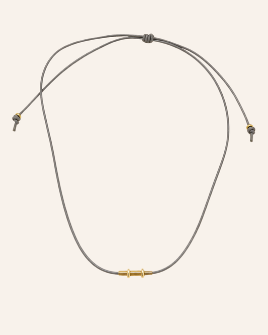 A minimalist adjustable cord necklace with a central gold-toned bead and gold-toned toggle details, displayed on a solid pale pink background.