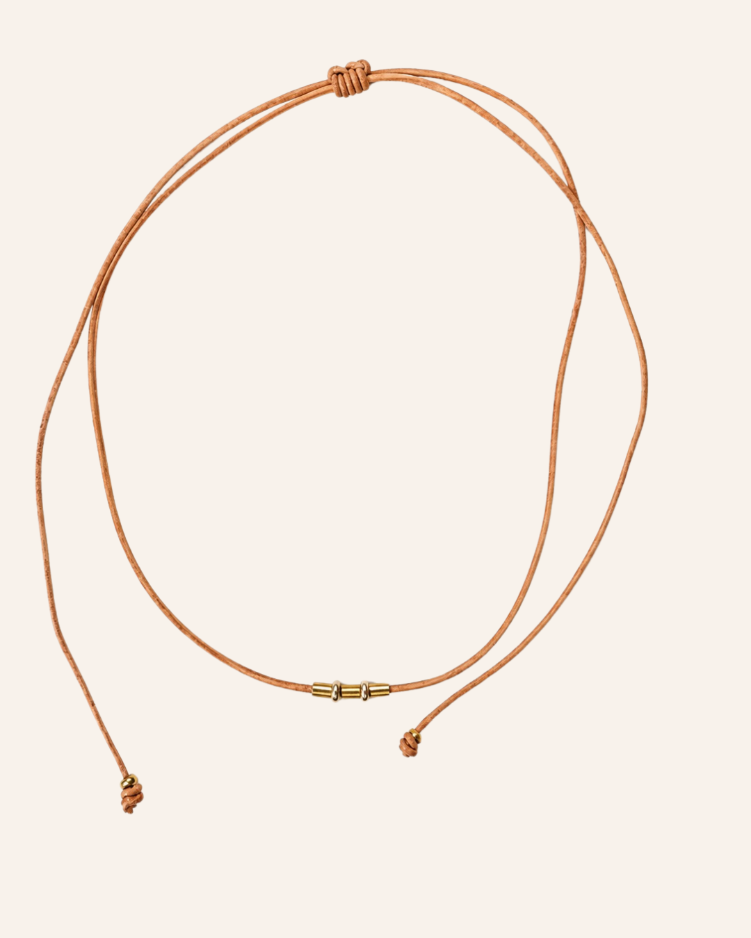 A double-strand adjustable cord necklace in a warm tan or cognac brown, featuring a small gold-toned metal connector in the center and knotted sliding ends, displayed on a solid pale beige background.