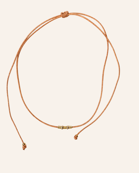 A double-strand adjustable cord necklace in a warm tan or cognac brown, featuring a small gold-toned metal connector in the center and knotted sliding ends, displayed on a solid pale beige background.