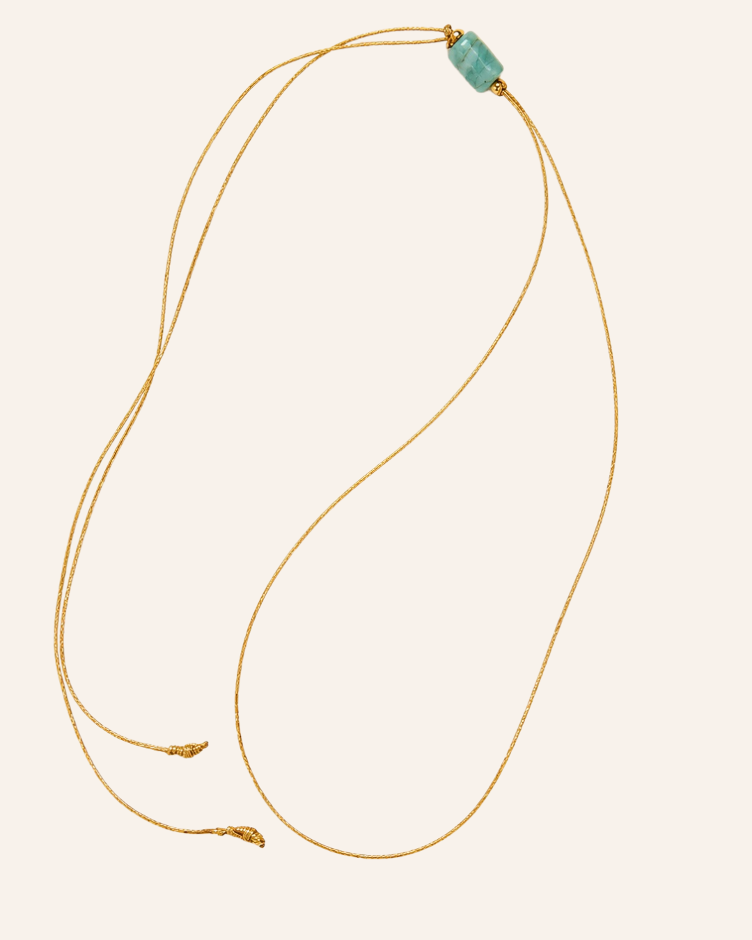 A delicate gold adjustable cord necklace featuring a small, polished light green stone bead at the top, displayed on a solid pale pink background.