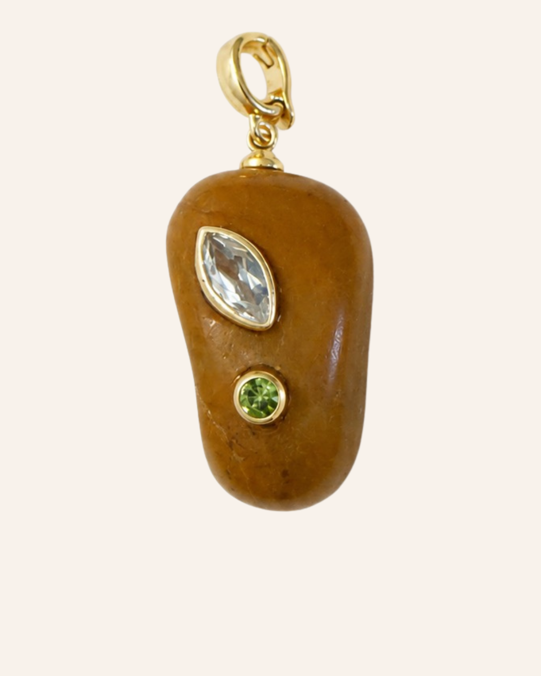 A gold pendant charm shaped like a smooth brown stone, featuring a marquise-cut clear gemstone and a small round green gemstone, set in gold on a light cream background.