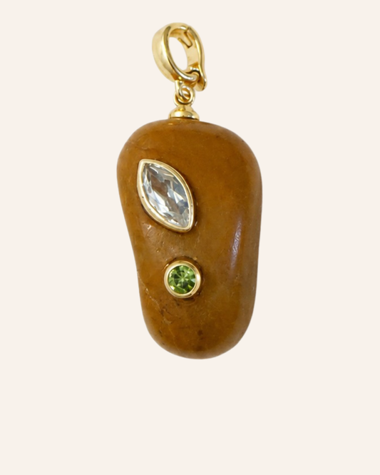 A gold pendant charm shaped like a smooth brown stone, featuring a marquise-cut clear gemstone and a small round green gemstone, set in gold on a light cream background.