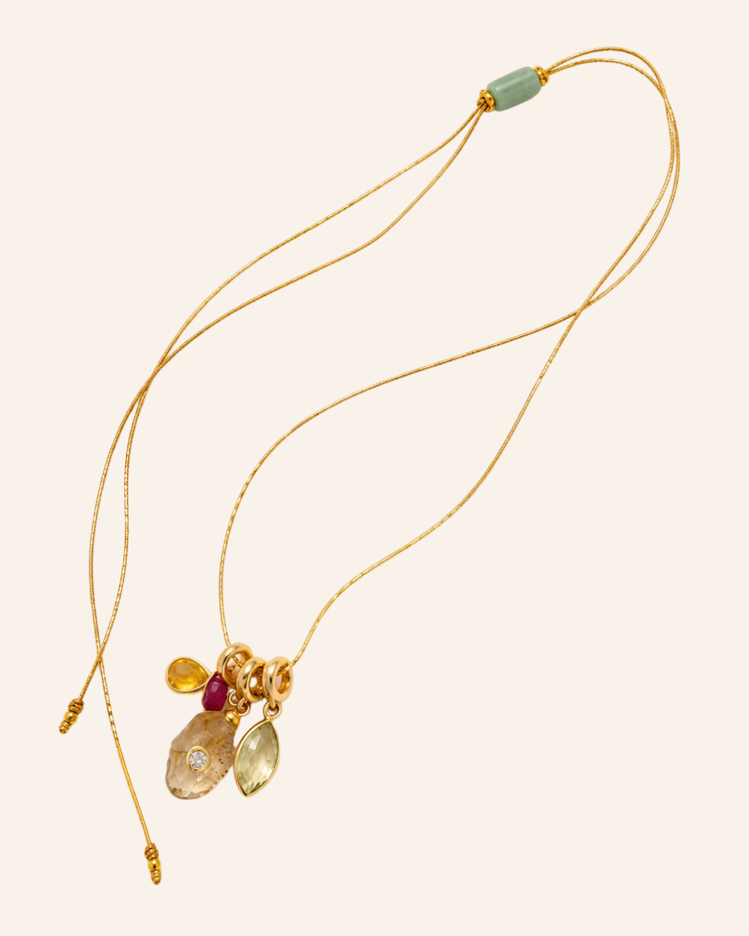 A delicate gold chain necklace with a multi-stone pendant featuring a light green bead, a yellow gemstone, a deep red gemstone, and a larger pale green gemstone, displayed on a solid pale pink background.