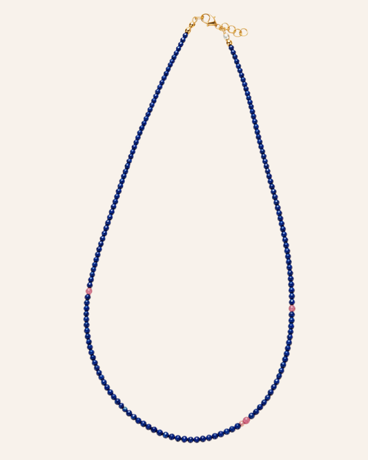 A delicate beaded necklace featuring deep blue faceted beads with small pink accent beads spaced along the strand, finished with a gold clasp and extender chain, set against a solid cream background.
