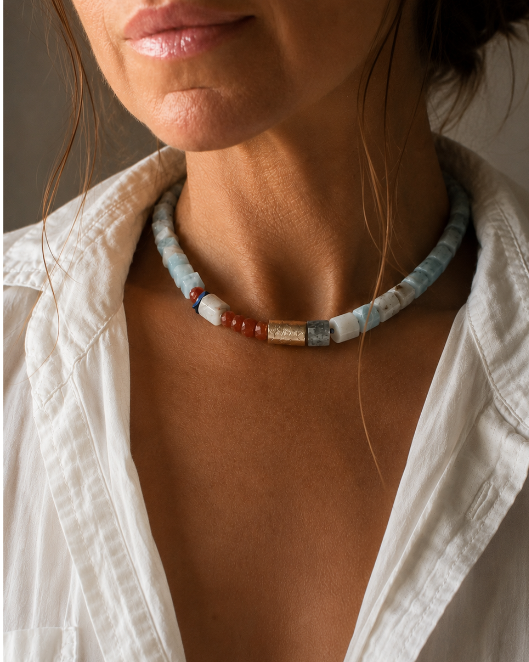 A close-up of a woman wearing a handmade beaded necklace made of light blue, white, and reddish-brown stones, paired with an open white shirt.