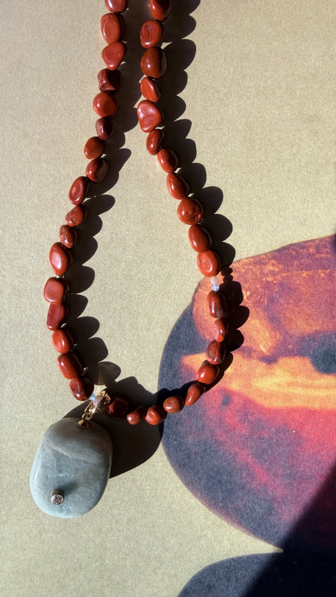 A beaded necklace made of reddish-brown polished stones with a smooth gray stone pendant, photographed on a tan surface with strong sunlight and shadows.